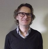 A white man with brown wavy hair wearing a black jumper and glasses